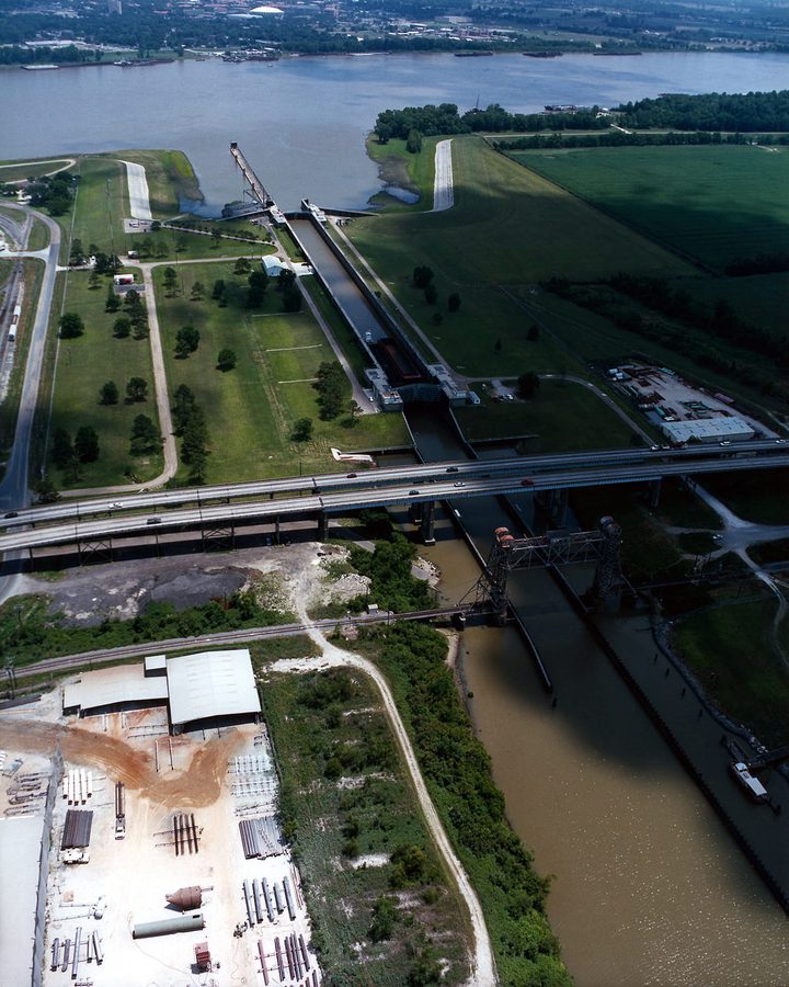 Aerial view of a Louisiana navigation lock where an industrial canal meets a wide river, crossed by a highway bridge and a railroad lift bridge, with a pipe yard in the foreground.